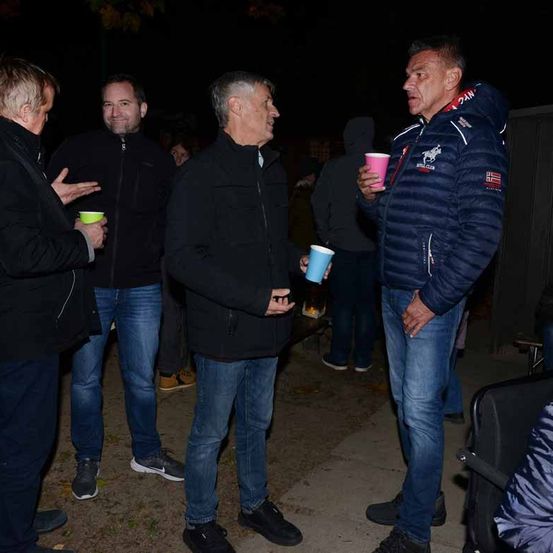 Eine Gruppe von Männern unterhält sich in einer nächtlichen Umgebung. Ein Mann hält einen rosa Becher, während ein anderer Mann einen blauen Becher hat. Der Mann auf der rechten Seite trägt einen blauen Mantel mit Logo.