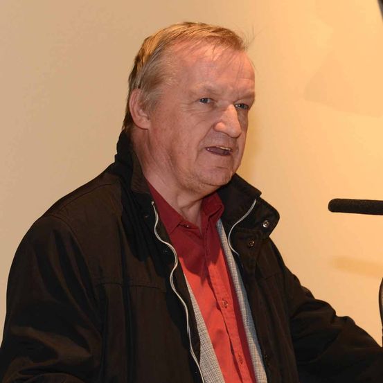 Ein älterer Mann mit kurzen blonden Haaren trägt eine schwarze Jacke und ein rotes Hemd. Er sitzt vor einem Mikrofon.