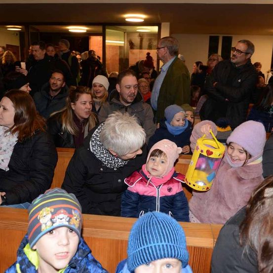 Eine große Menschenmenge in geschlossenen Räumen, einige in Winterkleidung, mit Kindern, die bunte Spielzeuge halten, und Erwachsenen, die auf Bänken stehen oder sitzen.