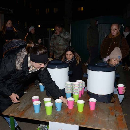 Eine Gruppe von Menschen steht um einen Tisch mit Bechern herum, einige mit Getränken darin, unter einem dunklen Himmel.
