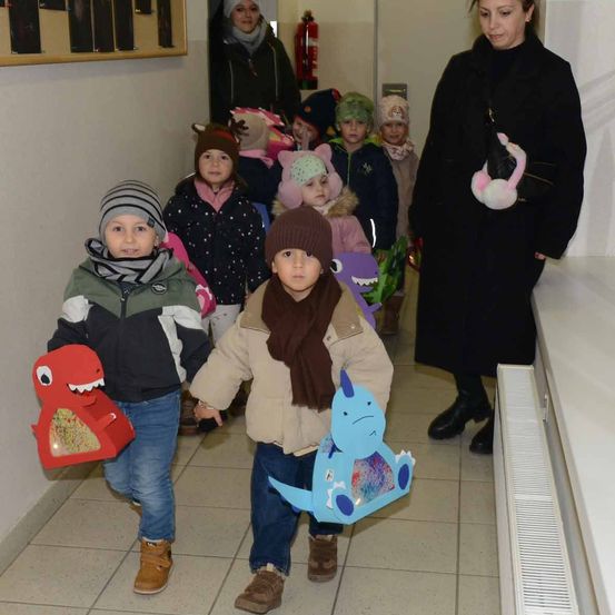 Mehrere Kinder in Winterkleidung, manche mit Masken, gehen durch einen Korridor. Ein Kind hält eine Dinosauriermaske, und ein Erwachsener folgt dahinter.