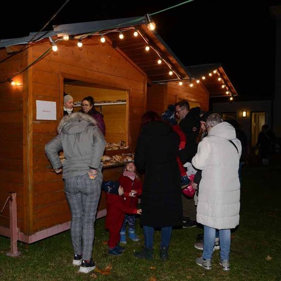 Mehrere Erwachsene und Kinder stehen in einer Schlange vor einem kleinen Holzstand, der Essen verkauft, der von Lichterketten beleuchtet wird, bei Nacht.
