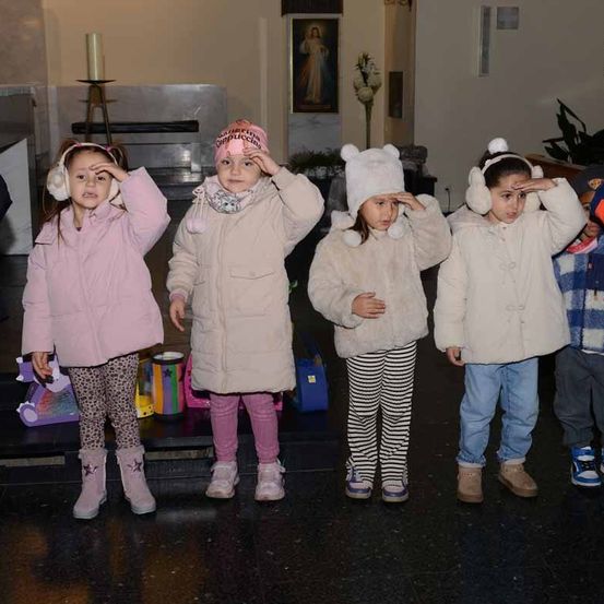 Eine Gruppe von Kindern in Winterkleidung steht in einer Kirche. Ein Mädchen trägt eine rosa Mütze, und andere haben Winterzubehör. Ein religiöses Bild ist im Hintergrund.