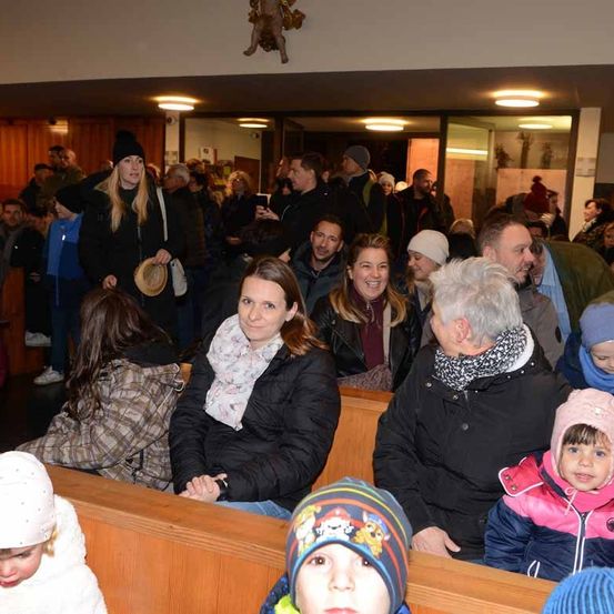 Eine große Gruppe von Menschen ist drinnen versammelt, viele tragen Winterkleidung. Sie sitzen auf Bänken, stehen und unterhalten sich. Eine Frau im Vordergrund hält einen Hut.