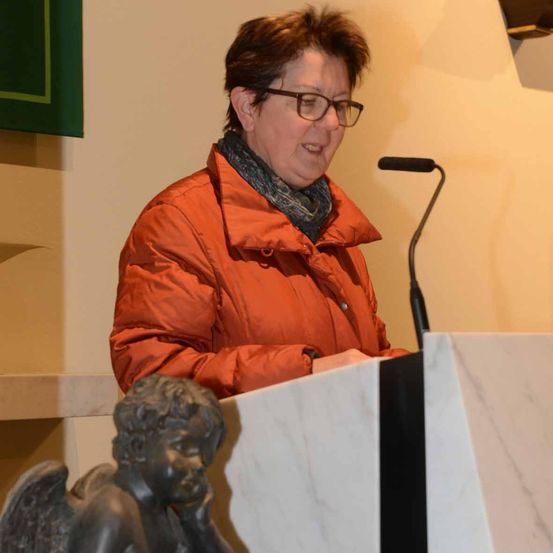 Eine Frau mit Brille und einer orangefarbenen Jacke steht an einem Rednerpult und spricht in ein Mikrofon. Hinter ihr befindet sich eine weiße Wand mit einem grünen Banner auf der linken Seite.