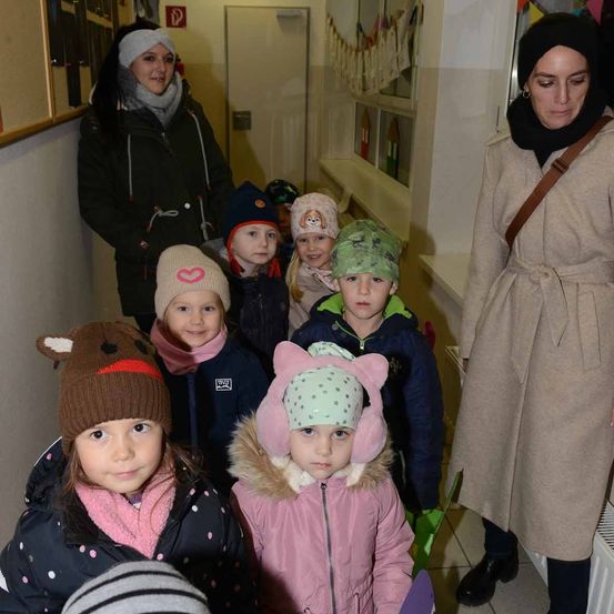 Eine Gruppe von Kindern in Wintermänteln und Mützen steht in einem Flur mit zwei Frauen. Eine Frau trägt einen Hijab und die andere eine Schürze. Hinter ihnen ist eine weiße Wand mit einem Fenster.