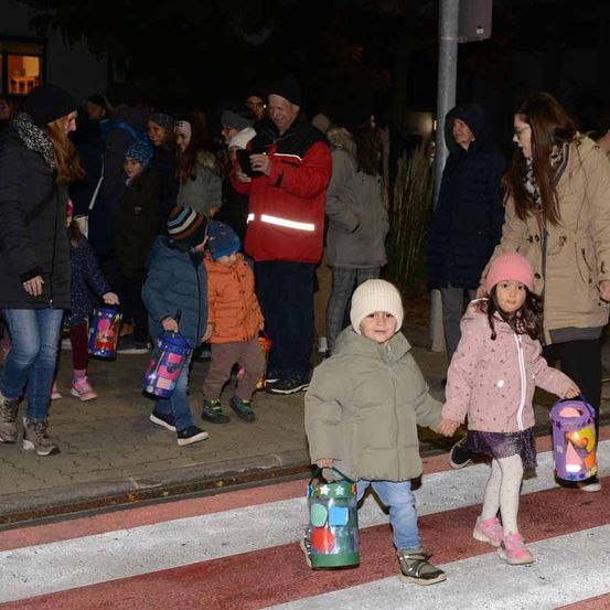 Eine Gruppe von Menschen, darunter Kinder, läuft über einen Zebrastreifen. Sie halten bunte Taschen. Die Umgebung scheint nachts zu sein.