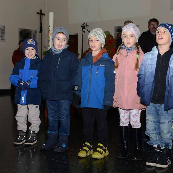 Eine Gruppe junger Kinder in Wintermänteln und Mützen steht zusammen in einer Kirche, möglicherweise singen sie. Hinter ihnen sind auch Erwachsene anwesend.