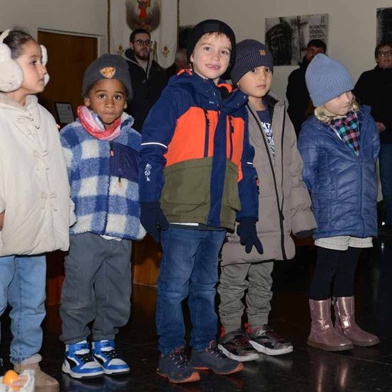 Fünf Kinder in Winterkleidung stehen drinnen zusammen. Zwei tragen Mützen, eines mit Schal und ein anderes trägt Stiefel. Dahinter stehen Erwachsene in der Nähe einer Wand mit gerahmten Bildern.