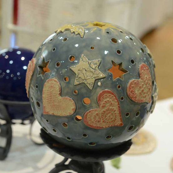 Eine handgefertigte Keramikkugel mit mehreren Herz- und Sternendesigns wird auf einem schwarzen Ständer ausgestellt. Die Kugel hat kleine Löcher und eine blaue Basis. Im Hintergrund ist eine weitere blaue Kugel zu sehen.