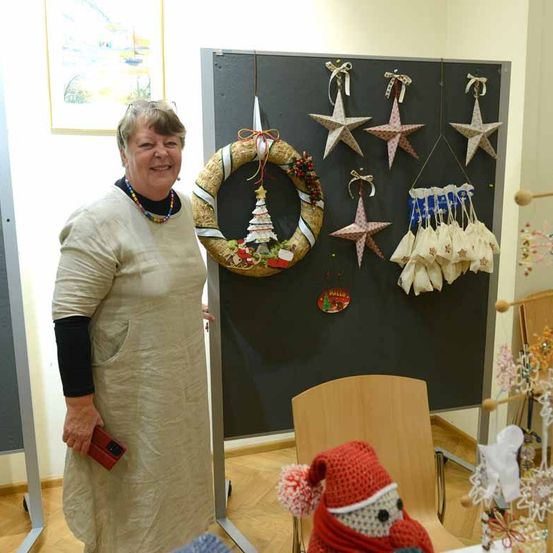 Eine Frau steht neben einer dekorierten Wand mit Weihnachtsdekorationen, einem Kranz und Sternen. Sie hält eine rote Geldbörse. Ein Holzstuhl und ein gehäkelter Schneemann sind in der Nähe.
