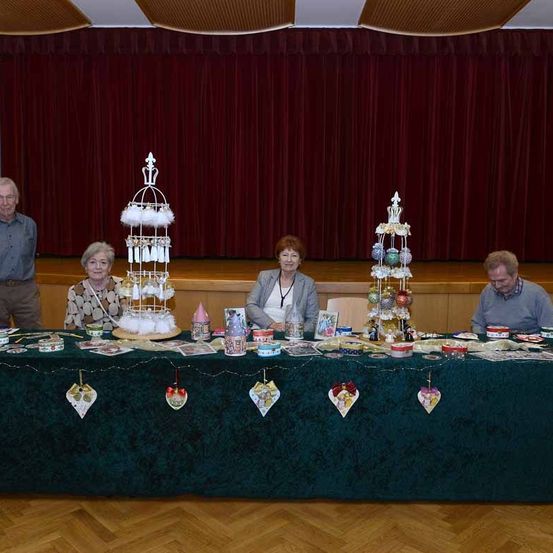 Vier Personen stehen hinter einem mit einem grünen Tuch bedeckten Tisch, der mit Weihnachtsdekorationen geschmückt ist. Zwei Frauen sitzen am Tisch, und zwei Männer stehen hinter ihnen. Der Tisch ist mit verschiedenen Ornamenten, Tassen und Karten verziert.