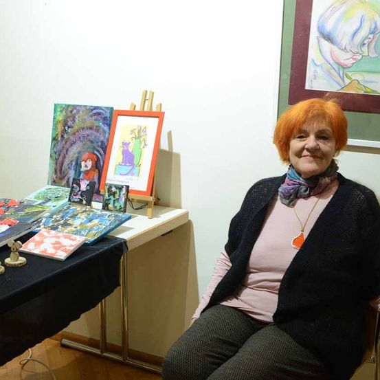 Eine Frau mit orangefarbenem Haar sitzt vor einem Tisch mit verschiedenen Kunstwerken. Der Tisch hat Gemälde, einen Rahmen und andere Kunststücke. Hinter ihr befindet sich eine weiße Wand mit einem gerahmten Gemälde.