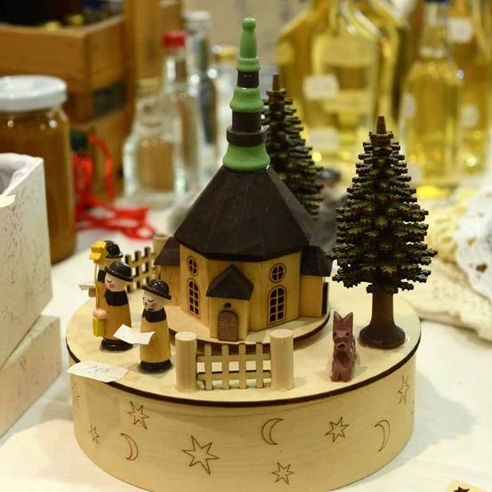 Eine hölzerne Krippe mit einer kleinen Kirche, zwei Figuren, einem kleinen Zaun und einem Baum. Dahinter sind Flaschen und Gläser im Hintergrund verschwommen.