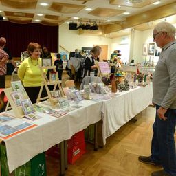 Ein Mann mit Brille und eine Frau in einem gelben Pullover stehen an einem Tisch mit verschiedenen ausgestellten Artikeln. Andere Leute sind verstreut, einige shoppen. Der Raum hat Holzböden und eine hohe Decke mit Lichtern.