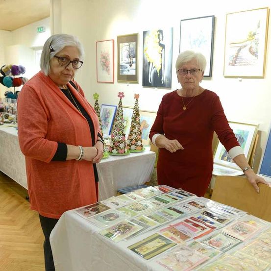 Zwei ältere Frauen stehen neben einem Tisch voller verschiedener Karten. Sie befinden sich in einem Raum mit gerahmten Gemälden an der Wand und einem Tisch mit einer weißen Tischdecke.