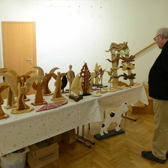 Ein Mann steht in einem Raum und beobachtet einen Tisch mit Holzskulpturen, darunter ein Hund. Der Tisch ist mit einer weißen Tischdecke bedeckt. Unter dem Tisch befinden sich Lagerboxen. Im Hintergrund sind eine Holztür und eine Treppe zu sehen.
