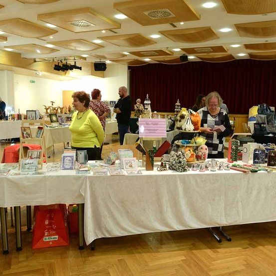 Ein Raum mit Holzboden und Deckenleuchten zeigt verschiedene Gegenstände auf einem Tisch, an dem Leute die Waren betrachten. Eine Frau in einem gelben Shirt steht am Tisch.