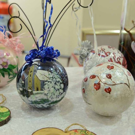 Mehrere handgefertigte Dekorationskugeln sind auf einem Tisch ausgestellt. Eine zeigt eine Winterlandschaft mit einem Haus und Schnee, eine andere zeigt einen Zweig mit roten Beeren.