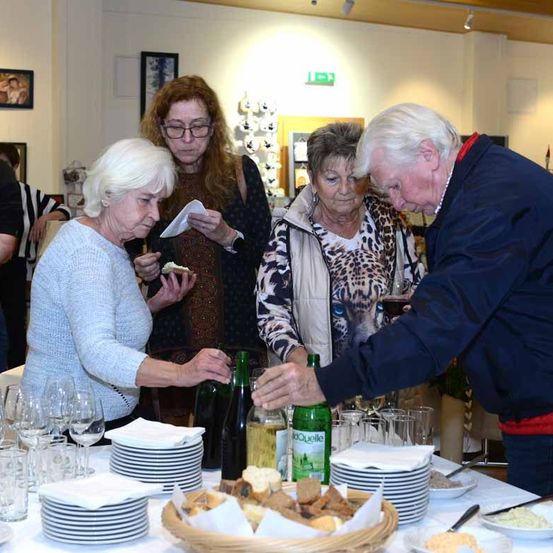 Eine Gruppe von Menschen sitzt an einem Tisch und genießt Essen und Getränke. Ein Mann schenkt Wein in Gläser, während andere essen und plaudern. Der Tisch ist mit Tellern, Weingläsern und verschiedenen Speisen gefüllt.