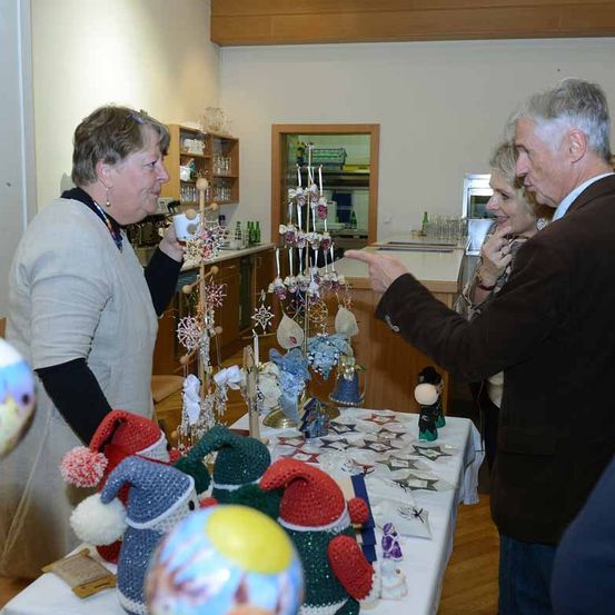 Eine ältere Frau steht hinter einem Tisch voller handgefertigter Gegenstände, lächelt und hält eine Tasse. Ein älterer Mann und eine Frau betrachten die Gegenstände, wobei der Mann darauf zeigt. Der Raum hat Holzwände und Regale mit Flaschen.
