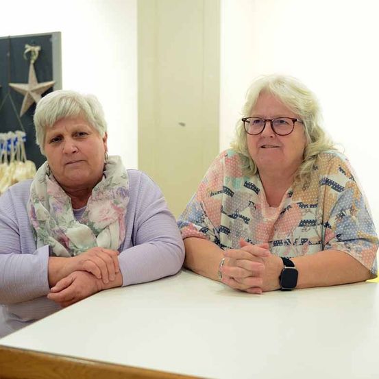 Zwei Frauen sitzen an einem Tisch, eine mit einem Schal und die andere mit einer Brille, beide lächeln. Hinter ihnen ist eine Wand mit Sternen und einer Tür.