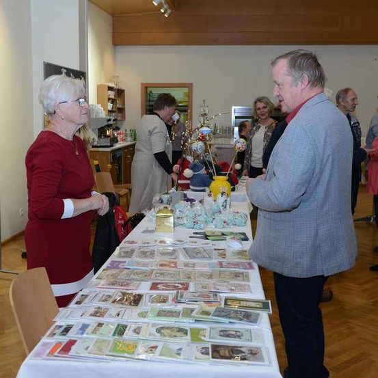 Eine Gruppe von Menschen befindet sich in einem Raum mit einem Tisch voller Karten und Geschenke. Eine ältere Frau und ein Mann unterhalten sich. Andere Leute stehen und betrachten die Gegenstände auf dem Tisch.
