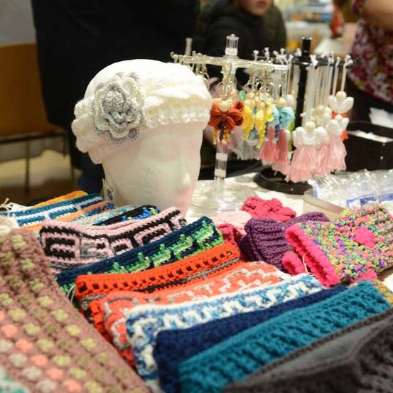 Eine Ausstellung von gehäkelten Accessoires, einschließlich Kopfbänder, Ohrringe und Handschuhe, steht auf einem Tisch. Ein Mannequinkopf mit einem gehäkelten Stirnband mit einer Blume sitzt auf dem Tisch.