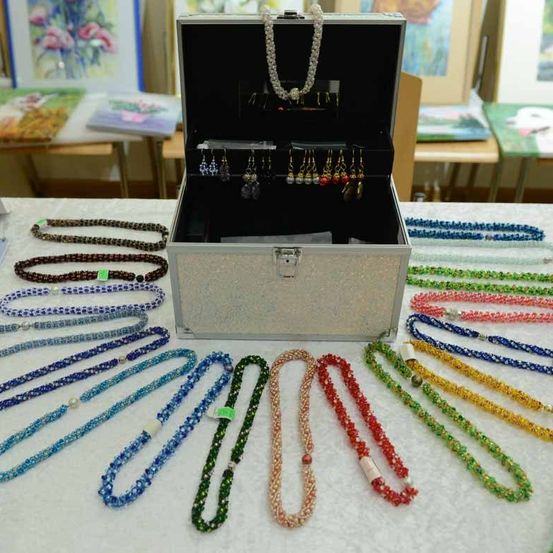 Eine Ausstellung verschiedener Halsketten und Ohrringe in einer silbernen Schatulle. Die Schatulle ist geöffnet und zeigt Fächer mit Schmuck darin. Zahlreiche Halsketten in verschiedenen Farben sind auf einer weißen Oberfläche ausgestellt.