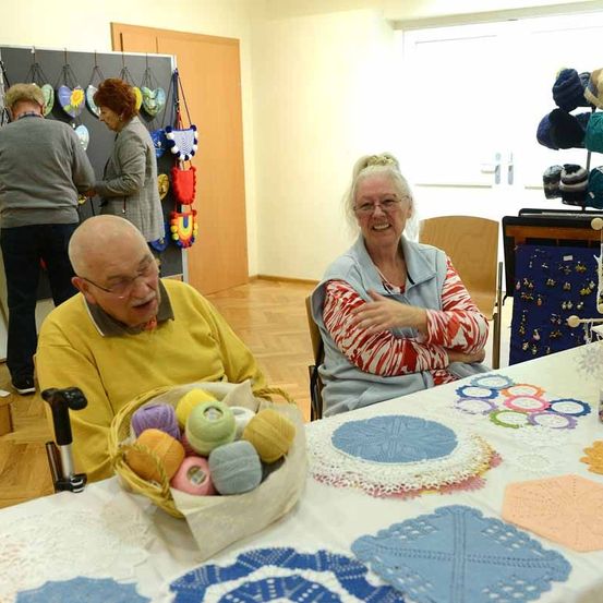Eine Gruppe von Menschen, einschließlich eines älteren Paares, sitzt in einem Raum mit Holzboden. Das ältere Paar sitzt vor einem Tisch mit Häkelarbeiten. Der Mann hat einen Korb mit Garn, und die Frau lächelt. Hinter ihnen befindet sich eine Wand mit einer hängenden Ausstellung von Häkelarbeiten.