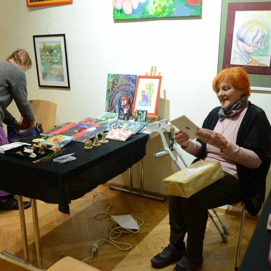 Eine Frau sitzt mit einem Geschenkkarton auf dem Schoß und lächelt in die Kamera. Hinter ihr steht ein Tisch, der mit Kunstutensilien und gerahmten Gemälden beladen ist. Eine andere Person steht in der Nähe des Tisches.