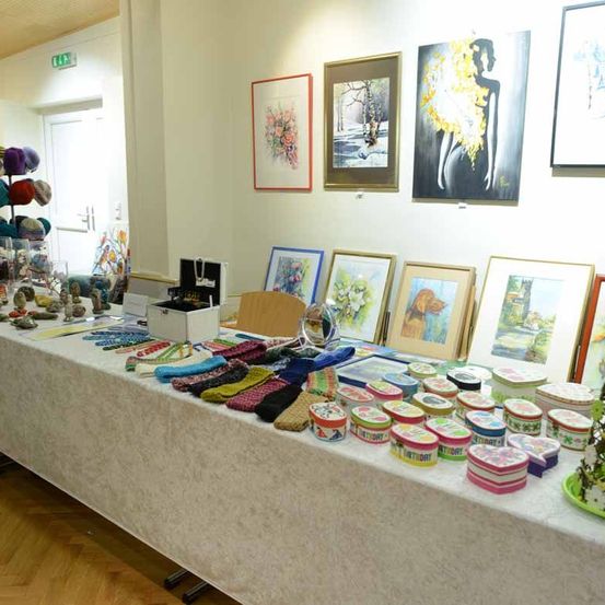 Ein Raum mit einem Ausstellungstisch, der mit verschiedenen Handwerksarbeiten und Gemälden bedeckt ist. Der Tisch ist mit bunten Schachteln, gestrickten Gegenständen und dekorativen Objekten bestückt. Gerahmte Gemälde hängen an der Wand darüber.