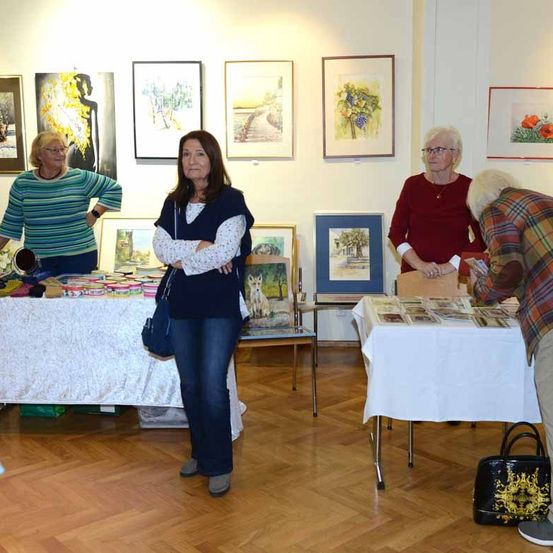 Vier Frauen sind in einer Kunstgalerie und betrachten Gemälde an der Wand. Zwei Tische haben Artikel zum Verkauf.