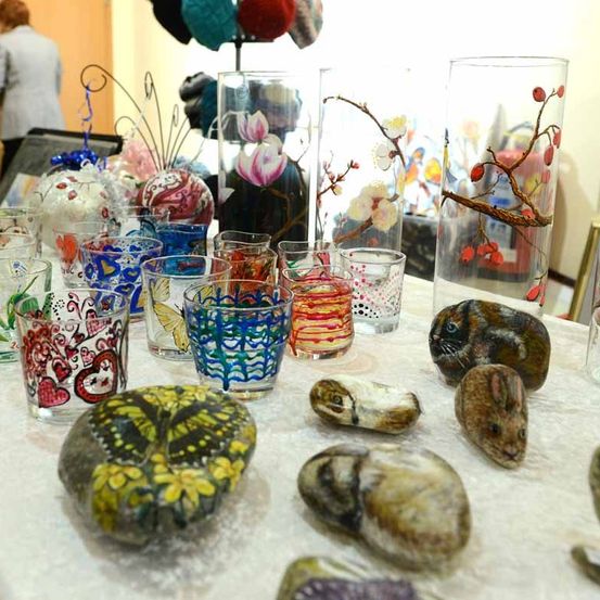 Ein Tisch ist mit einem weißen Tuch bedeckt und hat viele Gegenstände darauf. Bunte Gläser und Vasen sind angeordnet. Auf dem Tisch befinden sich bemalte Steine mit Tieren darauf.