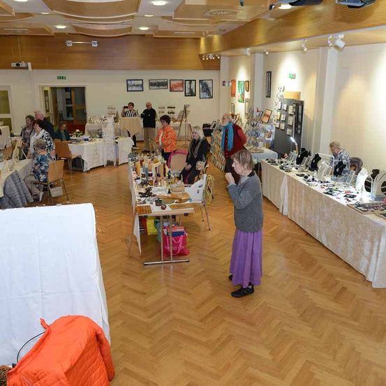 Ein Raum mit Holzböden und weißen Wänden beherbergt eine Versammlung von Menschen, die verschiedene Stände durchstöbern. Eine Frau in einer lila Schürze steht in der Mitte.