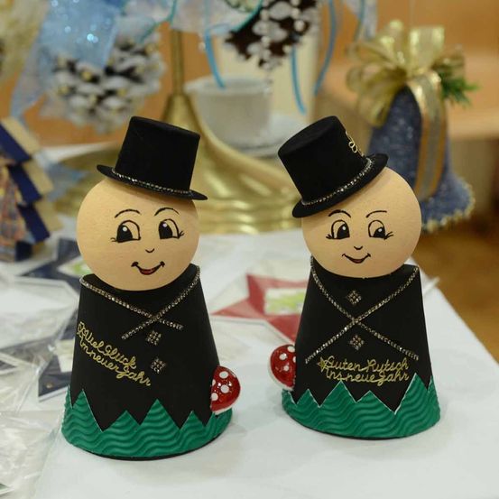 Zwei handgefertigte Figuren mit Zylinderhüten und lächelnden Gesichtern. Eine hat einen roten Pilz an der Basis und die andere einen roten Pilz auf der Schulter. Beide haben 'Guten Rutsch ins neue Jahr' darauf geschrieben. Dahinter befinden sich weitere Dekorationen und Ornamente.