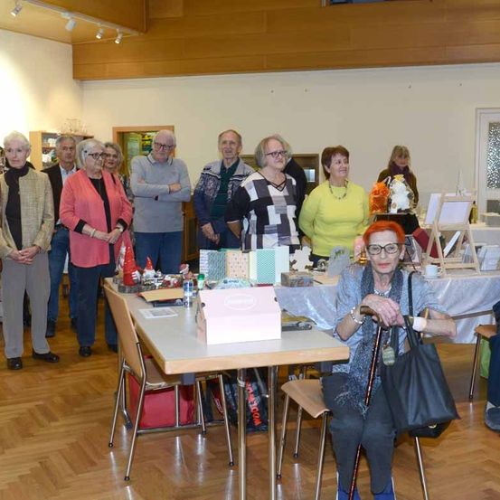 Eine Gruppe älterer Personen steht und sitzt in einem Raum, wobei eine Frau vorne einen Stock hält und eine Brille trägt. Hinter ihr befinden sich Tische mit verschiedenen ausgestellten Gegenständen, darunter eine rosa Schachtel und ein roter Gnom.