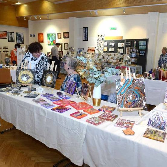 Ein Raum mit Holzböden beherbergt eine Kunstausstellung. Viele Gemälde und Kunstwerke sind auf Tischen ausgestellt. Besucher erkunden die Werke.