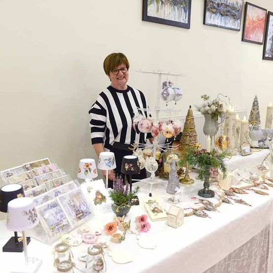 Eine Frau steht hinter einem Tisch, der mit Dekorationsartikeln wie Ornamenten, Blumen und Kerzen gefüllt ist. Der Tisch ist mit einem weißen Tuch bedeckt und sie trägt ein schwarz-weiß gestreiftes Hemd.