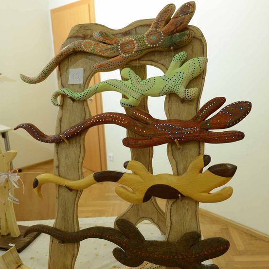Eine hölzerne Ausstellung zeigt geschnitzte Tierfiguren, darunter Eidechsen und Schlangen, die auf einer holzstruktur angeordnet sind. Die Figuren sind mit leuchtenden Farben und Mustern bemalt.