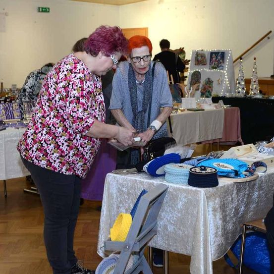 Zwei Frauen stehen vor einem Tisch mit gehäkelten Gegenständen, eine hält eine Geldbörse. Hinter ihnen steht eine weitere Person in der Nähe eines Tisches mit Kunstwerken. Der Raum hat andere Tische mit verschiedenen Gegenständen und eine Treppe mit einem Geländer.