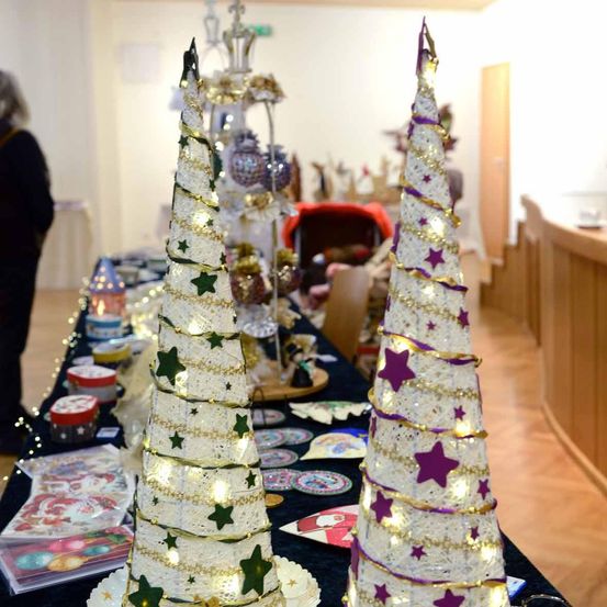 Ein Tisch zeigt zwei dekorative Weihnachtsbäume, einen mit grünen Sternen und Lichtern, den anderen mit lila Sternen und Lichtern. Verschiedene Ornamente und Lichter sind um die Bäume herum platziert. Eine Person steht im Hintergrund.