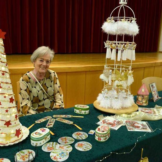 Eine ältere Frau sitzt hinter einem Tisch, der mit Weihnachtsdekorationen wie einem Weihnachtsbaum und einem Vogelkäfig-Ornament geschmückt ist. Die Umgebung ist in einem Raum mit einer Bühne und roten Vorhängen im Hintergrund.