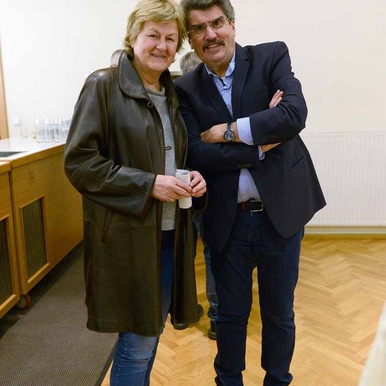 Eine Frau in einer braunen Lederjacke und ein Mann im Anzug stehen lächelnd zusammen. Sie halten eine Tasse und stehen in der Nähe einer Theke. Hinter ihnen steht eine Person in der Nähe eines Heizkörpers.