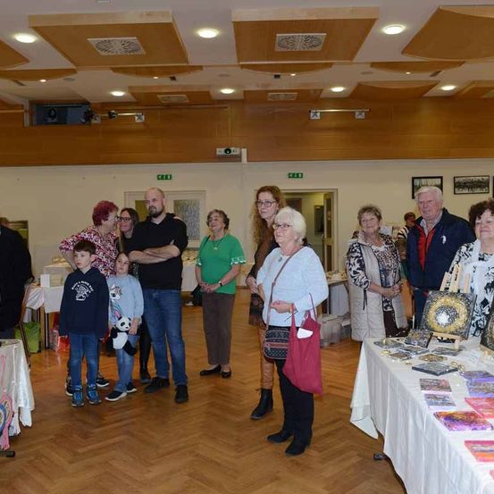 Mehrere Menschen stehen in einem Raum mit Holzböden und Tischen, auf denen verschiedene Gegenstände ausgestellt sind. Eine Frau hält eine rote Tasche. Ein Projektor und Lautsprecher sind an der Wand montiert.