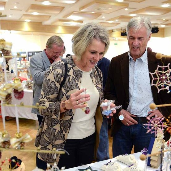 Eine Frau und ein Mann stehen neben einem Tisch mit Dekorationen und lächeln sich an. Hinter ihnen schaut ein Mann im Anzug nach unten. Der Raum hat Holzböden und Tische mit verschiedenen ausgestellten Artikeln.