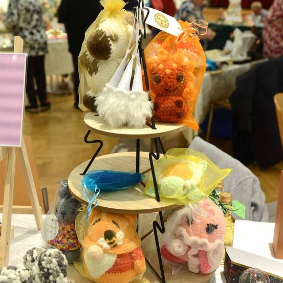 Ein dreistufiger Stand zeigt verschiedene gehäkelte Plüschtiere, darunter eine weiße Katze, einen orangefarbenen Fuchs und ein rosa Schwein. Diese sind in durchsichtigen Plastiktüten präsentiert. Im Hintergrund sind Menschen zu sehen, möglicherweise auf einem Markt.
