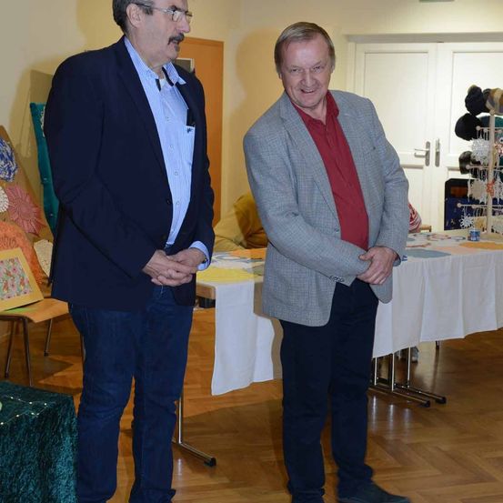 Zwei Männer stehen nebeneinander, lächelnd, in einem Raum mit Tisch und Holzboden. Hinter ihnen stehen Stühle, ein Tisch mit Gegenständen und eine Wand mit einer Tür.