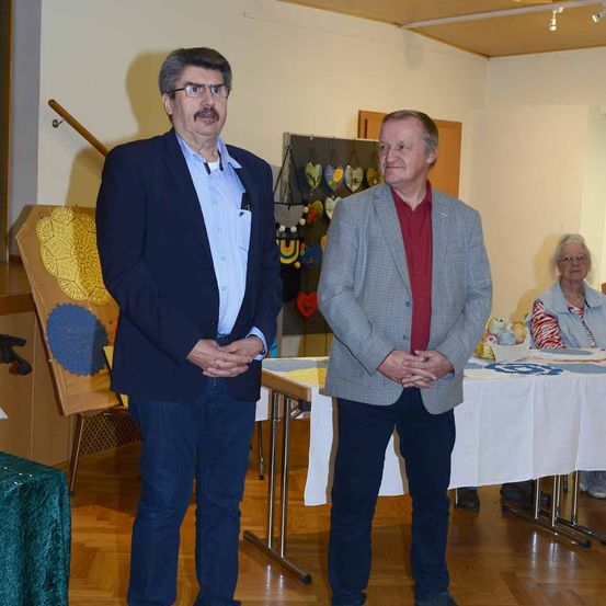 Zwei Männer stehen nebeneinander in einem Raum und tragen Geschäftskleidung. Hinter ihnen sitzt eine Frau an einem Tisch mit weißer Tischdecke. Verschiedene Gegenstände, darunter ein Holzstaffelei und ein Holzstuhl, befinden sich im Raum.