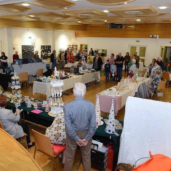 Ein großer Raum mit Holzböden und Tischen, die mit verschiedenen Gegenständen bedeckt sind, wahrscheinlich ein Markt oder ein Handwerksmarkt. Die Leute stöbern und einige stehen in einer Schlange.